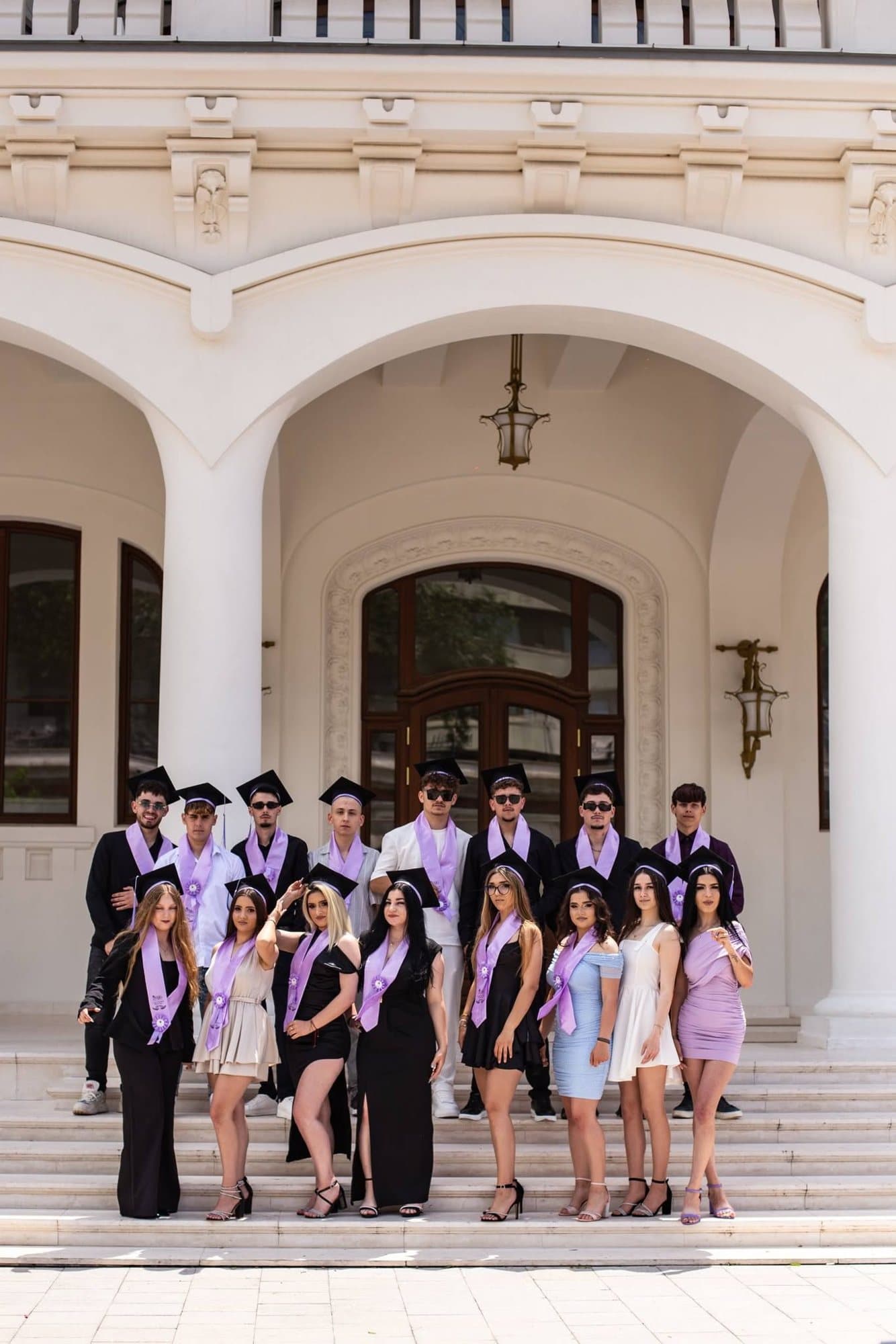 Fotografie de grup a unei clase de absolvenți cu eșarfe lila, pe treptele liceului